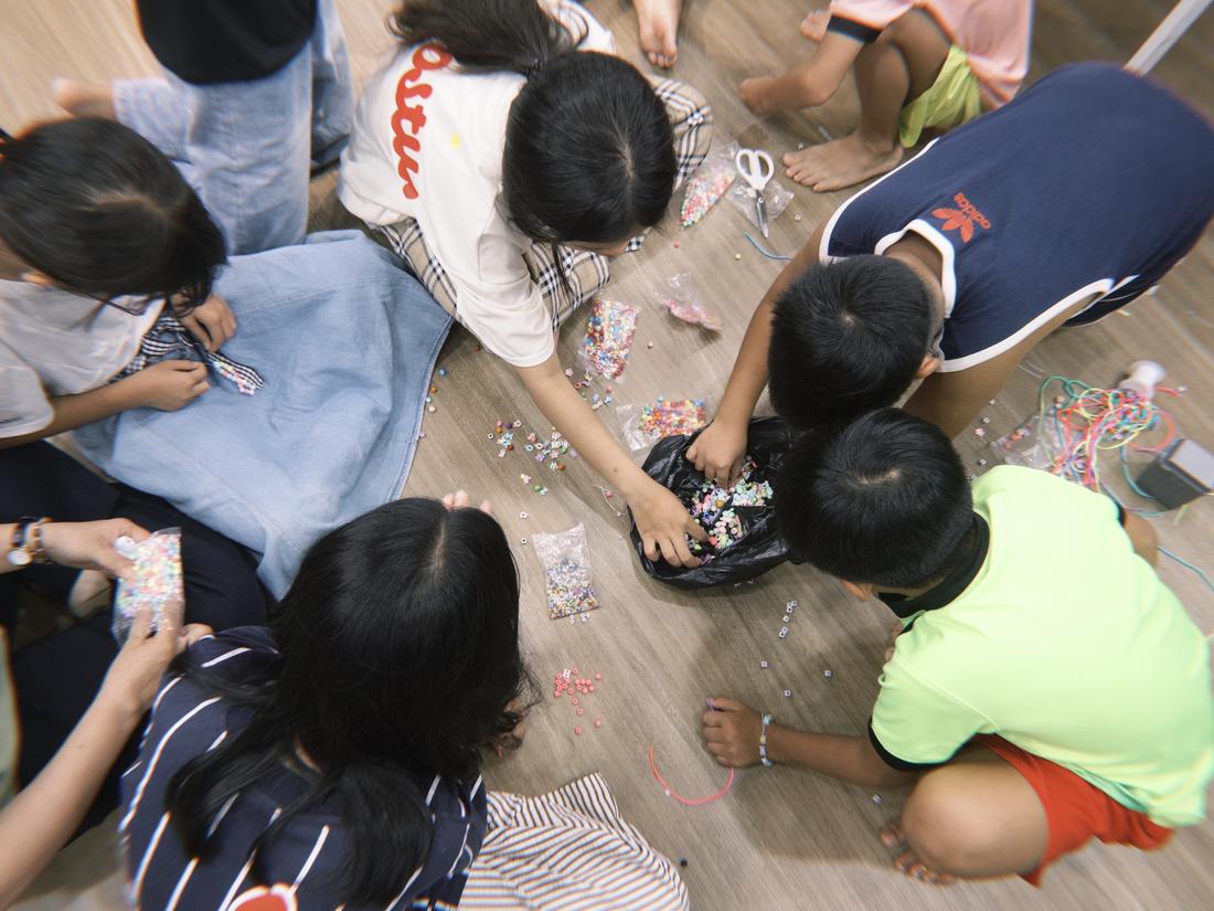 Overhead photo of volunteers and children working together on bracelets on the floor.